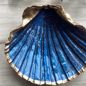 May include: A decorative seashell-shaped dish. The interior is a deep blue with gold speckles, and the rim is trimmed with gold. The dish has a glossy finish and is likely used for holding small items or as a decorative piece.