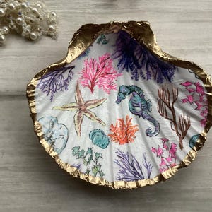 May include: A decorative seashell with a gold-painted rim, featuring a hand-painted underwater scene. The design includes a starfish, seahorse, coral, and seaweed in shades of pink, orange, blue, and purple.