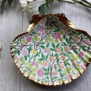May include: A decorative seashell-shaped dish with a gold-painted rim. The interior features a floral pattern with pink, yellow, and green flowers on a white background. The dish is placed on a light grey wooden surface.