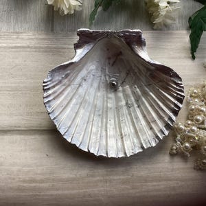May include: A silver-painted scallop shell-shaped dish with a small, round stud in the centre. The dish has a textured surface and is on a light-coloured wooden surface. A strand of pearls is to the right.