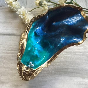 May include: An oyster shell dish with a gold-painted edge. The inside is filled with blue and teal resin and has three small pearl accents. The shell is on a light wooden surface, with white flowers in the background.