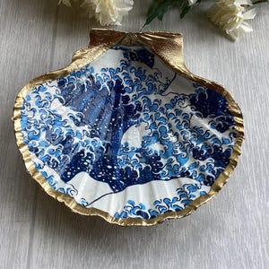 Peut inclure: Un plat en forme de coquille Saint-Jacques décoré d'un motif graphique bleu et blanc représentant des vagues stylisées. L'intérieur de la coquille présente ce motif, tandis que les bords extérieurs et la charnière sont peints en doré.