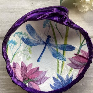 May include: A decorative seashell with a vibrant purple rim. The interior features a blue dragonfly and floral design in shades of blue, pink, and green. A unique, handcrafted item.