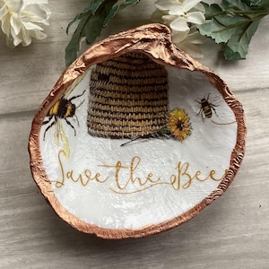 May include: A decorative oyster shell painted with a bee theme. The shell features a beehive, bees, and a yellow flower. The words "Save the Bee" are written in gold script. The shell's edge is trimmed with a metallic copper color.