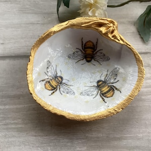 May include: A decorative seashell trinket dish with a gold-painted rim. The white interior features three illustrations of bees with black and yellow stripes. The dish is placed on a light-colored wooden surface, with floral accents.