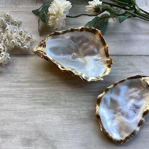 May include: Two oyster shell jewellery dishes with gold-painted edges. One shell holds a single pearl. A beaded pearl necklace and white flowers are also in the image. The shells are on a light grey wooden surface.