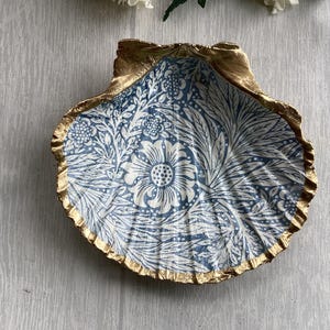 May include: A decorative scallop shell dish featuring a blue and white floral pattern on the interior surface. The scalloped edges of the shell are painted with a metallic gold finish, highlighting the natural ridges and texture of the shell.