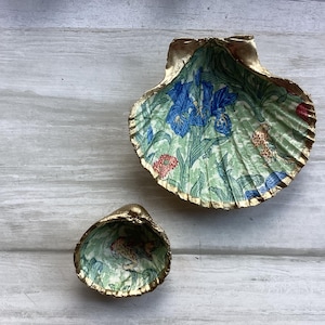 May include: Two decorative seashells with a gold trim. The larger seashell has a green, blue, and yellow floral pattern. The smaller seashell has a similar pattern with a bird.
