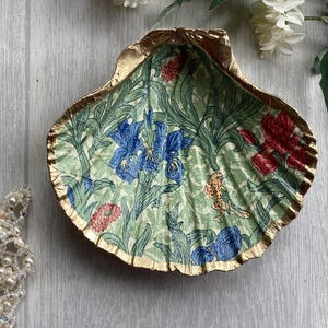 May include: A decorative seashell trinket dish with a gold-painted rim. The interior features a floral pattern with blue irises, red flowers, and a small bird on a light green background.