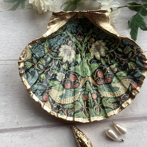 May include: A decorative seashell trinket dish with a gold-painted rim. The interior features a detailed floral and bird pattern in shades of green, cream, and red. The dish is a unique and artistic home decor item.