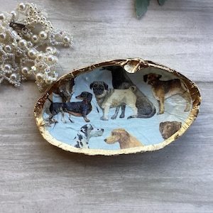 May include: A decorative seashell dish with a gold-painted rim. The interior features a collage of various dog breeds on a light blue background. A pearl necklace is visible in the background.