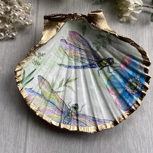 Peut inclure: Un coquillage Saint-Jacques décoratif orné d'un motif de deux libellules colorées et de fleurs bleues sur un fond vert pâle. Les bords festonnés du coquillage sont peints avec une finition dorée métallique, soulignant les stries naturelles.