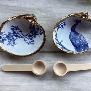May include: Two white ceramic seashells with gold trim. One shell has a blue and white floral design, the other has a blue peacock design. Two wooden spoons are also pictured.