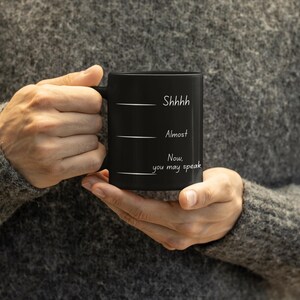 Person holding a black coffee mug printed with the phrase “Shhh… Almost… Now you may speak,” emphasizing the mug’s size and design.