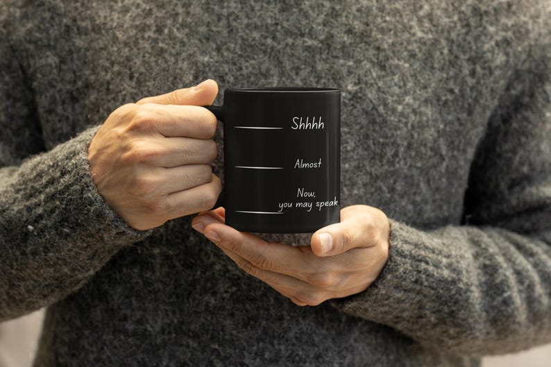 Person holding a black coffee mug printed with the phrase “Shhh… Almost… Now you may speak,” emphasizing the mug’s size and design.