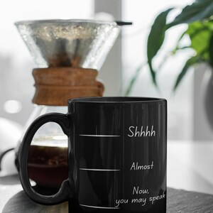 Black mug with humorous quote “Shhh… Almost… Now you may speak” next to a pour-over coffee setup on a kitchen counter.