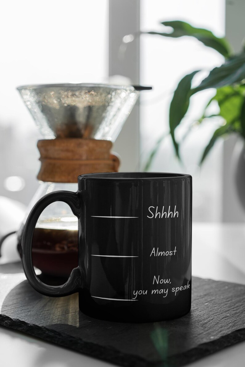 Black mug with humorous quote “Shhh… Almost… Now you may speak” next to a pour-over coffee setup on a kitchen counter.