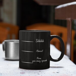 Black mug with white handwritten-style quote “Shhh… Almost… Now you may speak” resting on a kitchen counter near cookware.