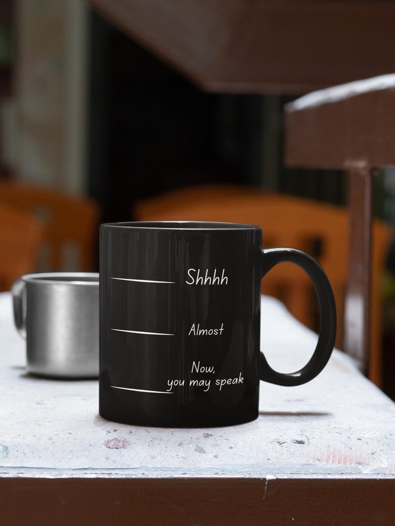 Black mug with white handwritten-style quote “Shhh… Almost… Now you may speak” resting on a kitchen counter near cookware.