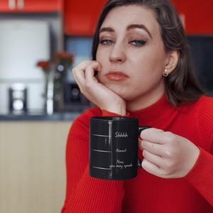 Woman sitting at a table holding a black mug with the quote “Shhh… Almost… Now you may speak,” styled as a morning coffee moment.
