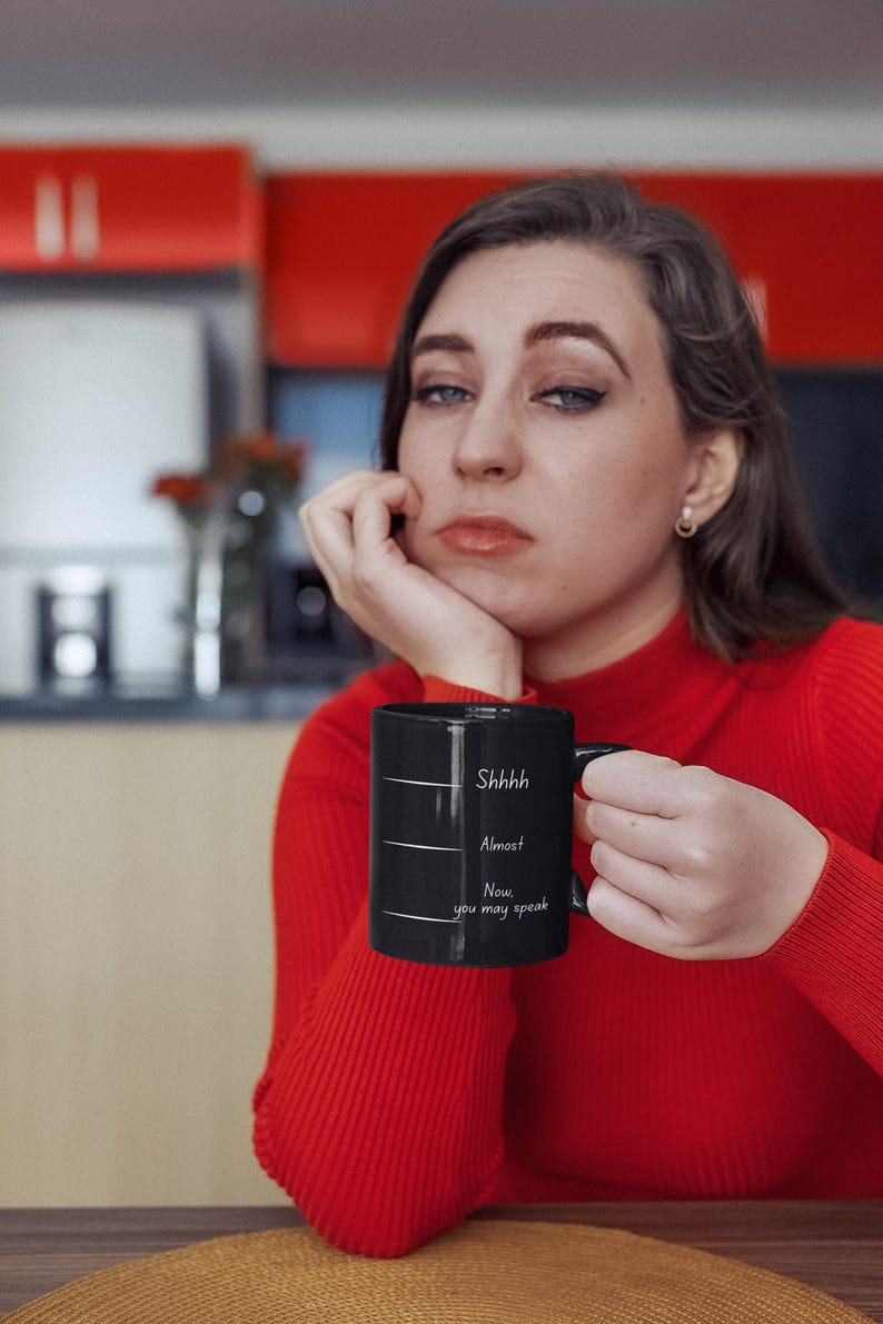 Woman sitting at a table holding a black mug with the quote “Shhh… Almost… Now you may speak,” styled as a morning coffee moment.