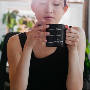 Woman holding a black coffee mug with “Shhh… Almost… Now you may speak,” conveying a quiet, reflective coffee break.