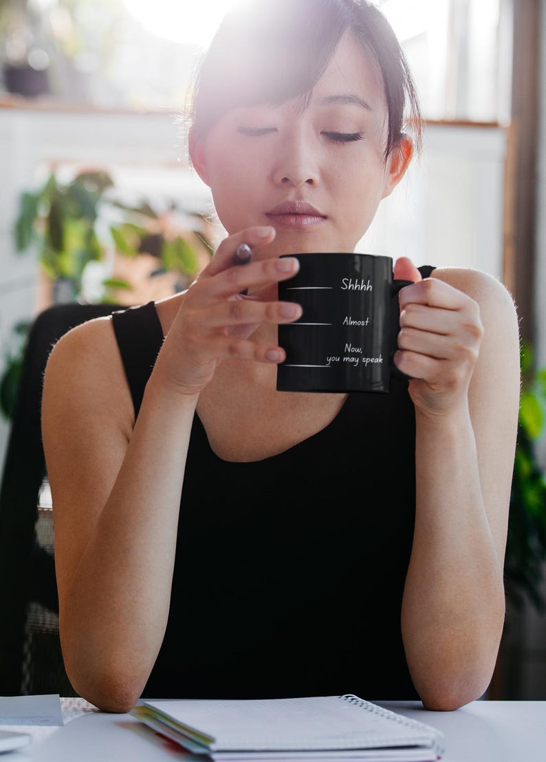 Woman holding a black coffee mug with “Shhh… Almost… Now you may speak,” conveying a quiet, reflective coffee break.