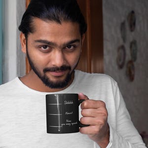 Man holding a black coffee mug featuring the phrase “Shhh… Almost… Now you may speak,” shown in a relaxed lifestyle portrait.