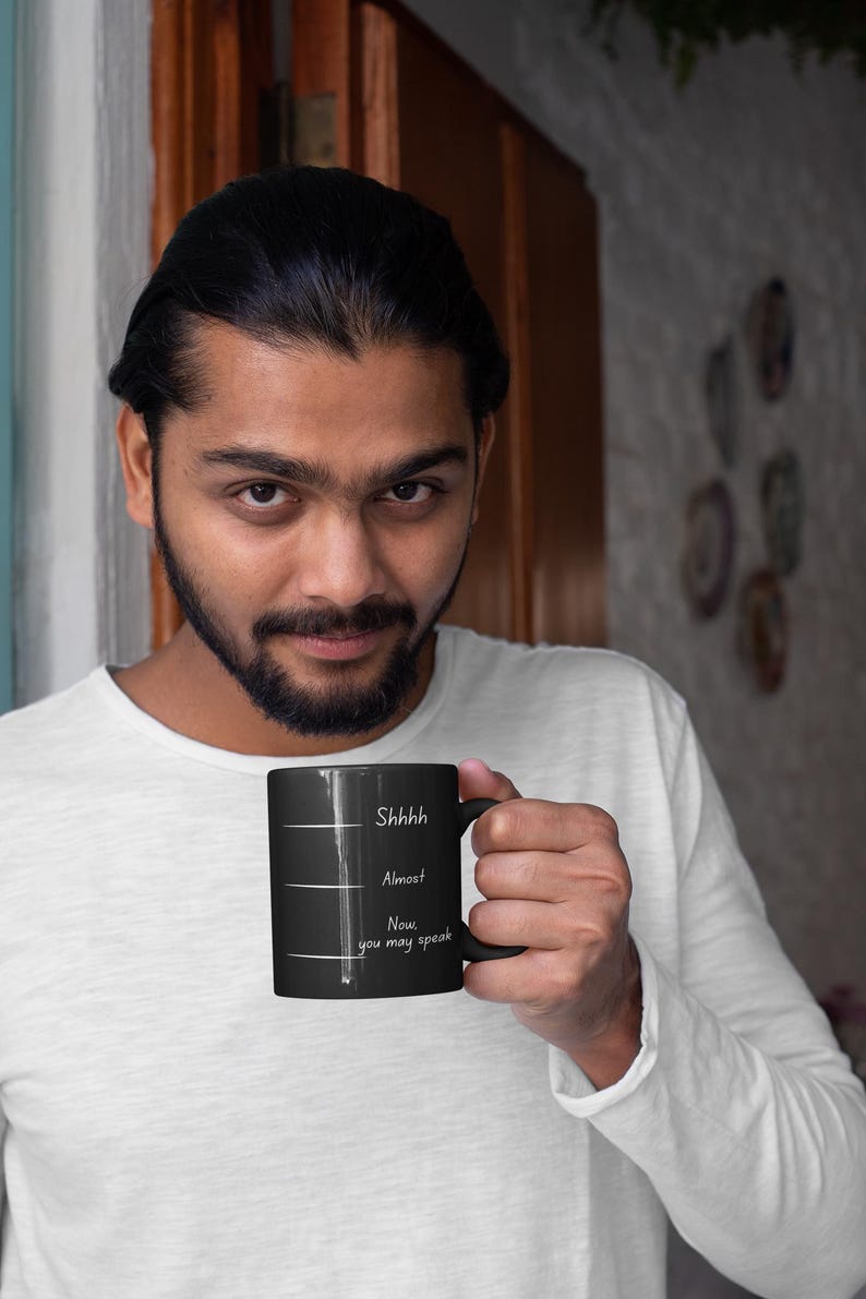 Man holding a black coffee mug featuring the phrase “Shhh… Almost… Now you may speak,” shown in a relaxed lifestyle portrait.