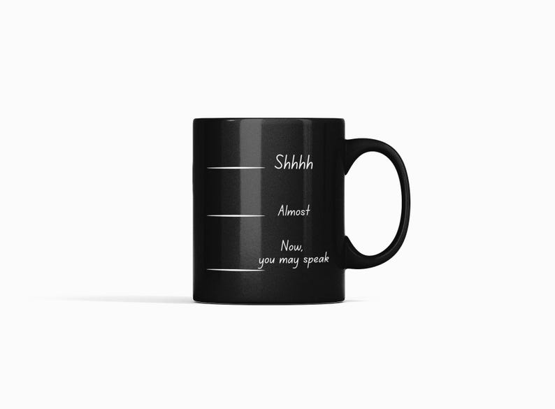 Black ceramic coffee mug with white handwritten-style text reading “Shhh… Almost… Now you may speak,” featuring a minimalist humorous design.