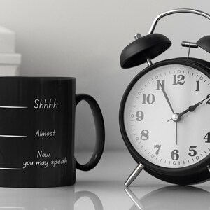 Black coffee mug with the phrase “Shhh… Almost… Now you may speak” placed beside a classic alarm clock, suggesting a morning routine.