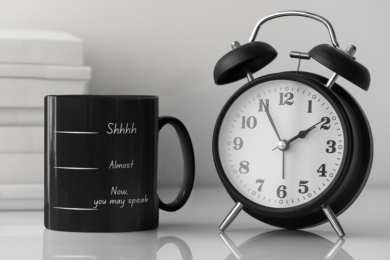 Black coffee mug with the phrase “Shhh… Almost… Now you may speak” placed beside a classic alarm clock, suggesting a morning routine.
