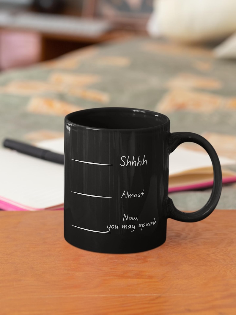 Black mug with humorous quote “Shhh… Almost… Now you may speak” placed on a wooden surface in a casual workspace setting.