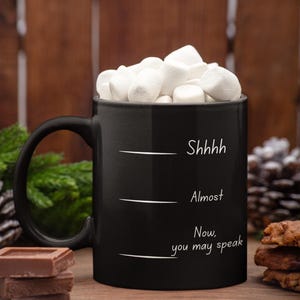 Black mug with “Shhh… Almost… Now you may speak” filled with marshmallows, styled in a cozy fall or winter setting.