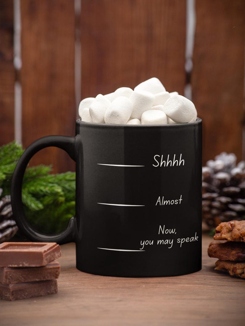 Black mug with “Shhh… Almost… Now you may speak” filled with marshmallows, styled in a cozy fall or winter setting.