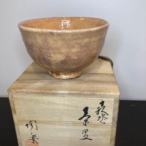Puede incluir: Un cuenco de té de cerámica marrón claro con una superficie texturizada, sentado sobre una caja de madera con caracteres japoneses escritos en ella. La caja tiene una tapa marrón oscura.