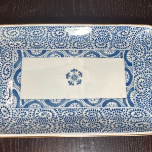Pode incluir: Um prato retangular de cerâmica branca com um padrão floral azul e branco. O prato tem uma borda dourada e uma seção central elevada.