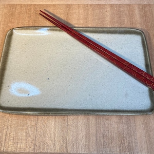 May include: A rectangular, light brown ceramic plate with a dark brown rim. Two red chopsticks with gold accents are placed on the plate.