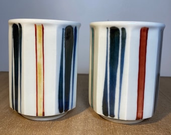 Puede incluir: Dos tazas de cerámica blanca con un diseño geométrico. Las tazas tienen rayas verticales en tonos de azul, verde, rojo y amarillo.