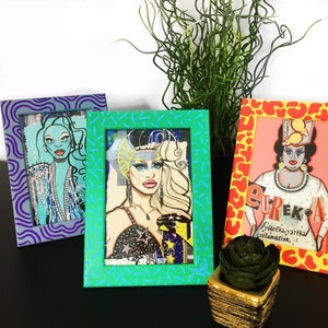 Puede incluir: Tres ilustraciones enmarcadas de drag queens. Los marcos est&aacute;n decorados con diferentes patrones: remolinos morados, confeti verde y azul, y estampado animal naranja y amarillo. Las ilustraciones presentan las caras y los nombres de las reinas. La primera reina lleva un atuendo azul y plateado y tiene el pelo azul. La segunda reina lleva un atuendo negro y plateado y tiene el pelo rubio. La tercera reina lleva un atuendo blanco y tiene el pelo casta&ntilde;o. El texto en el tercer marco dice "Eureka!" y "Your flaws, your reclamation."