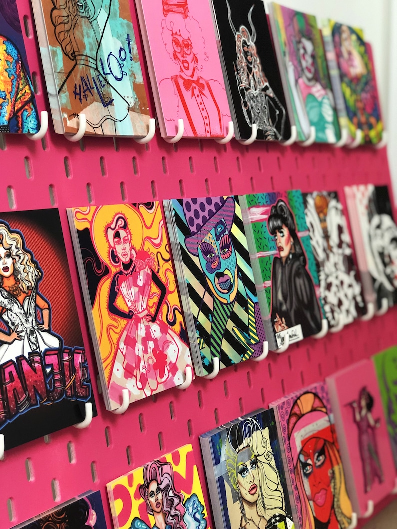 Puede incluir: Un tablero de clavijas rosa con ilustraciones coloridas de drag queens. Las ilustraciones est&aacute;n impresas en cartulina blanca y se cuelgan del tablero de clavijas con ganchos blancos. Las ilustraciones presentan a varias drag queens en diferentes poses y atuendos. Algunas de las ilustraciones incluyen texto, como "Hallelooot" y "Annie".