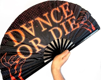 May include: Black hand fan with the words "DANCE OR DIE" in orange block letters. The fan has an orange dotted pattern and orange claw-like graphics. The fan is held open against a white background.