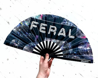 May include: A black folding fan with a disco ball pattern and the word "FERAL" in white block letters. The fan is held open, revealing a black and white handle. The fan is a unique accessory.