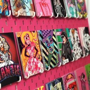 May include: A pink pegboard with colorful cards featuring illustrations of drag queens. The cards are arranged in rows and columns. Some of the cards have text on them, including "Hallelooot" and "Manila".