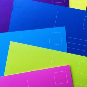 May include: A stack of colorful postcards with white dotted lines and a blank space for an address. The postcards are in shades of purple, blue, green, and pink.