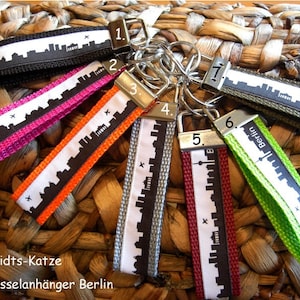 Könnte beinhalten: Zehn bunte Schlüsselanhänger mit schwarz-weißen Stadt-Skyline-Designs. Jeder Schlüsselanhänger hat eine Nummer und das Wort "Berlin" darauf gedruckt. Die Schlüsselanhänger sind in einem Kreis auf einer geflochtenen Weidenoberfläche angeordnet.