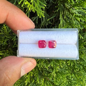 May include: Two cushion-cut, vibrant pinkish-red gemstones are displayed side-by-side in a clear, rectangular plastic storage box. The box features a white, soft, cylindrical foam insert designed to hold the two loose stones securely in place.
