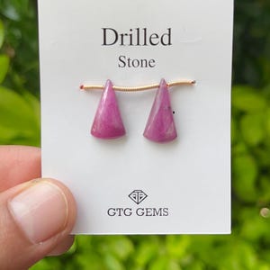 May include: Two pink triangle-shaped stones with holes drilled through them. The stones are strung on a gold wire and are labeled "Drilled Stone" and "GTG Gems".