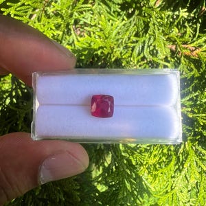 May include: A cushion-cut, deep red gemstone displayed in a clear plastic box with a white velvet insert. The gemstone features a faceted surface that reflects light, showcasing its vibrant color and geometric shape within the protective container.