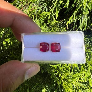 May include: Two cushion-cut, deep red gemstones are displayed side-by-side in a clear, rectangular plastic storage box. The box features a white, soft foam insert with two horizontal slots designed to hold the loose, faceted red stones securely.